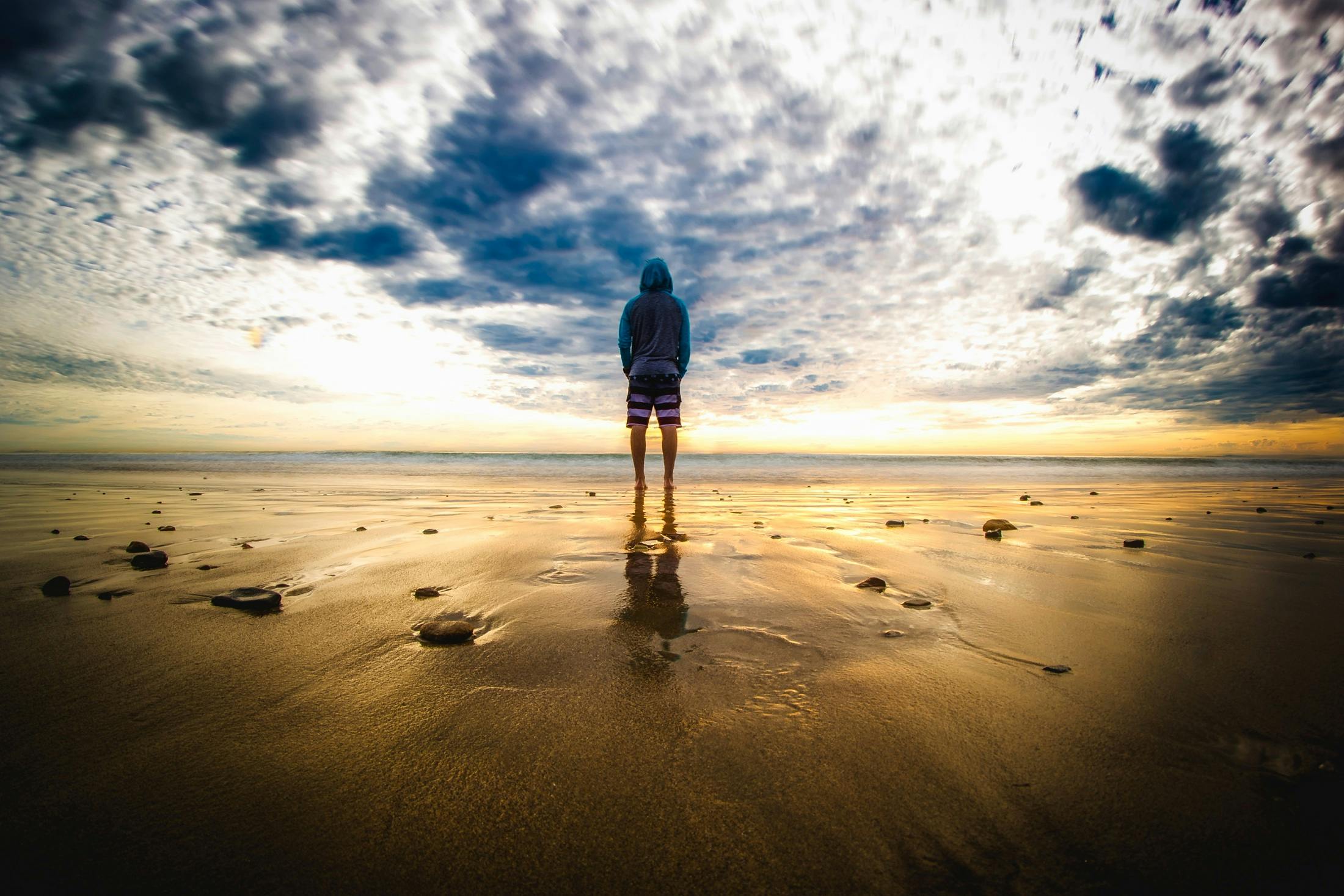 A lone figure stands on a tranquil beach during a vivid sunset, reflecting on the wet sand.