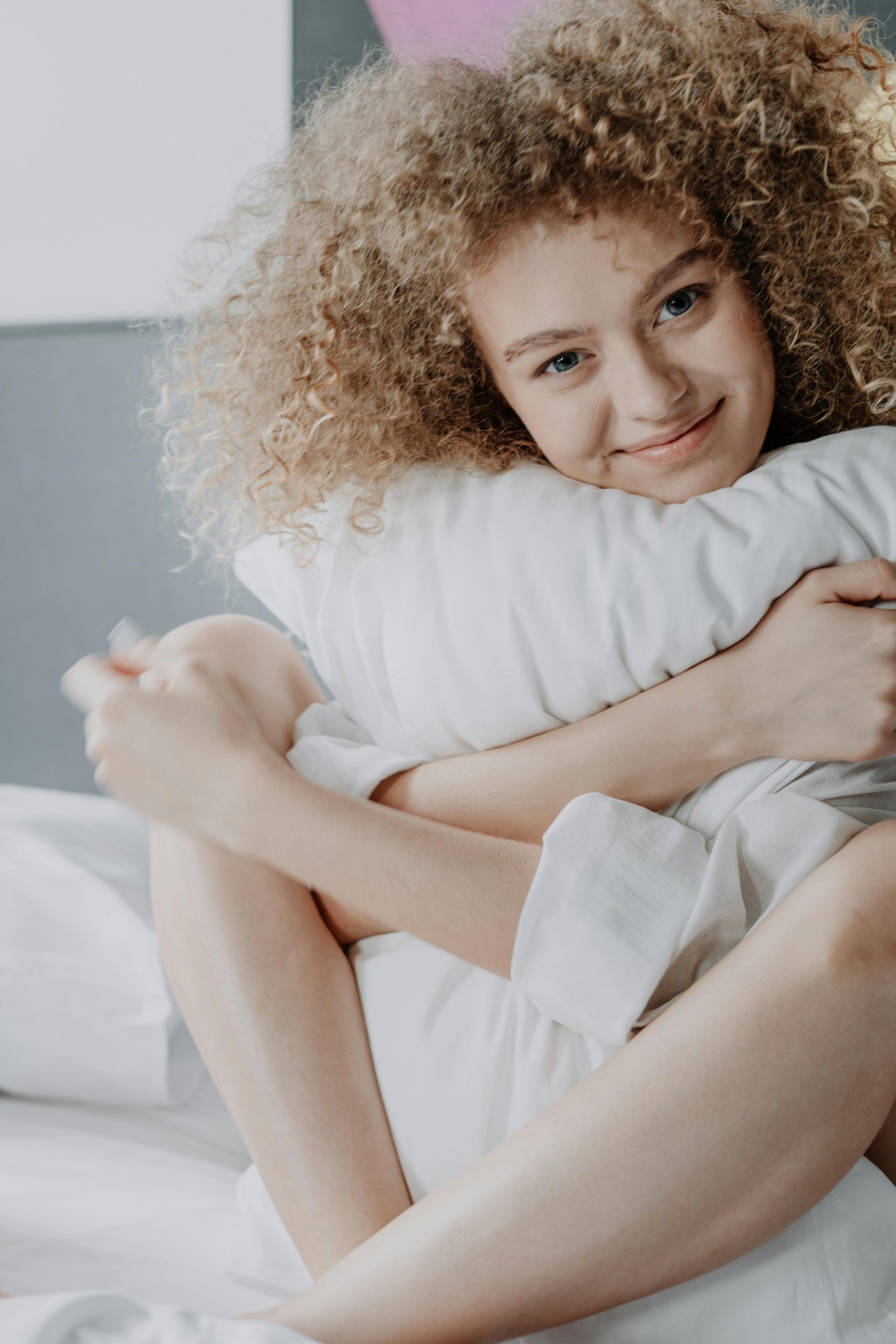 Joyful woman with curly hair hugging a pillow while smiling in a cozy bedroom setting.