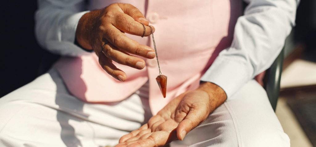 A man in soft pastel attire holding a gemstone pendulum over his palm outdoors.