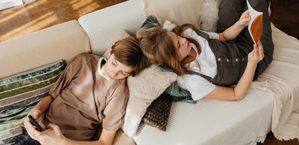 A boy and girl relax on a couch, reading and using a smartphone at home during the day.